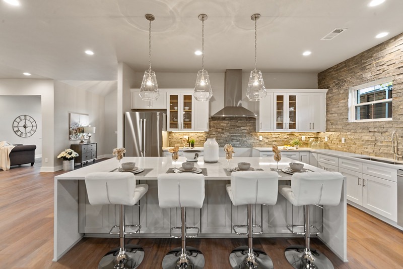 Modern kitchen featuring a white island with bar stools, pendant lights, and granite countertops. Stone accents and large windows add elegance.